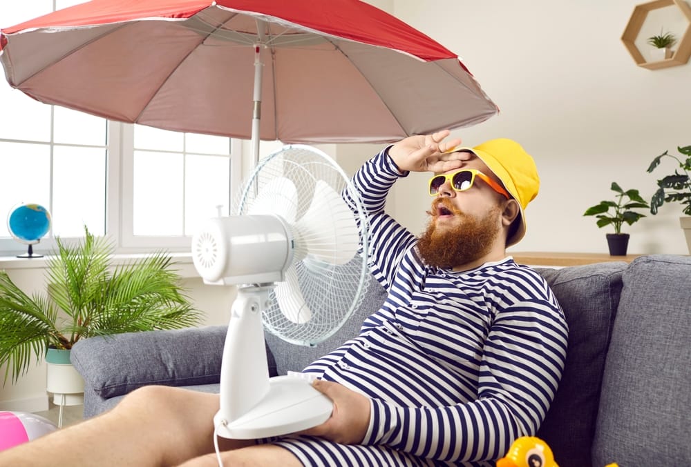 A person in a striped outfit and yellow hat sits on a couch under an umbrella, using a fan and appearing hot or tired, as if waiting for an auto draft to cool things down.