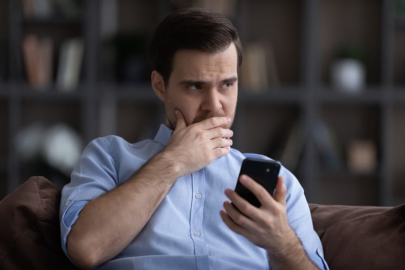 A man in a blue shirt is sitting on a couch, holding a smartphone and looking concerned with his hand covering his mouth. A blurred bookshelf is in the background, possibly hinting at an auto draft message causing his anxiety.